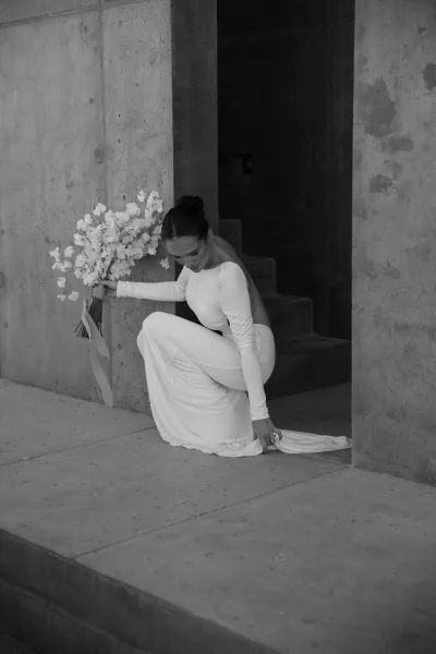 Bridal portrait of a bride adjusting her dress train beside a concrete wall and steps near a stark doorway.