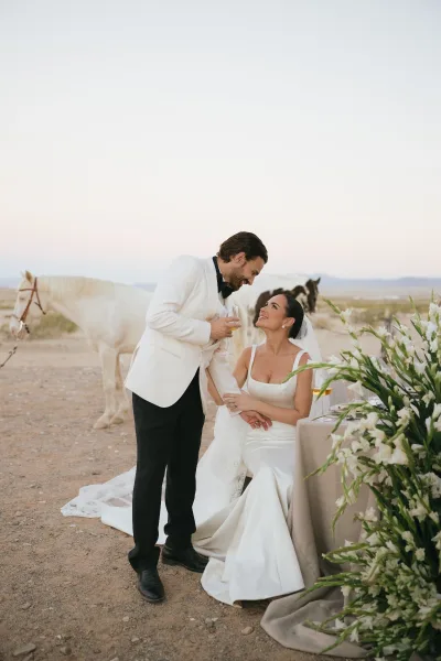 Couple portrait capturing a sweetheart table moment as newlyweds toast with champagne glasses against a desert landscape with horses.