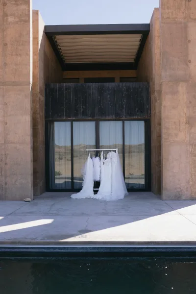 Bridal gowns lined up, a wedding dress on hanger beside a feather boa, framed by sliding glass doors to a desert pool patio.