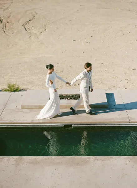 Couple portrait of bride and groom walking hand in hand along a poolside concrete walkway, the bride in a bridal gown, set against desert landscape.