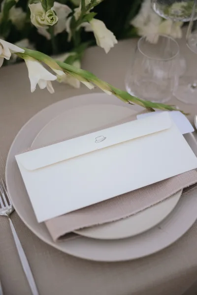 Wedding place setting in a minimalist table setting with an envelope resting on the plate, arranged on a tabletop with greenery.