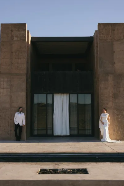 Couple portrait of a bride and groom standing apart, groom in a white tuxedo, set against a concrete building with glass doors and desert hills.