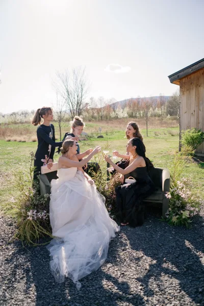 Bridesmaid toast as bride with bridesmaids raise champagne flutes on a loveseat, outdoors by gravel path and hills under blue sky