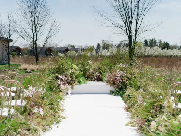 Ceremony aisle decor with outdoor wedding aisle lined by floral arrangements and greenery, white aisle runner, bentwood chairs in a meadow field under blue sky