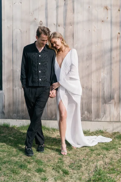 Couple portrait of bride and groom holding hands, her pleated long-sleeve thigh-slit gown beside his black suit by a wood plank wall outdoors