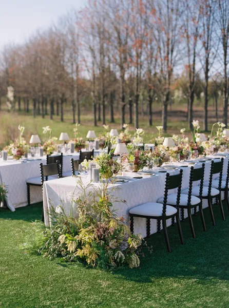 Reception tablescape at an outdoor wedding reception with long banquet tables, white linens, floral centerpieces, and lamps on a grassy lawn