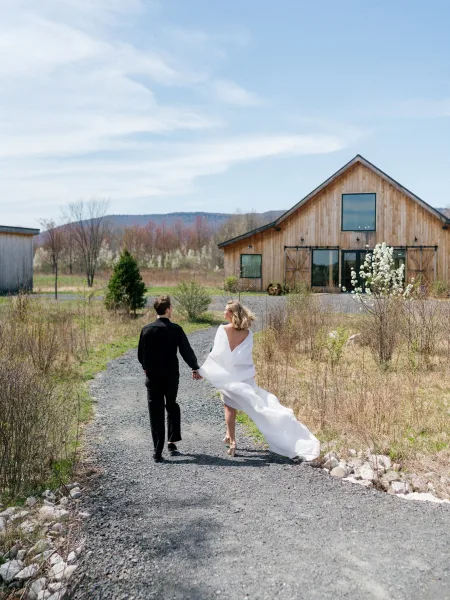 Couple portrait of bride and groom walking away holding hands, bride looking back as her cape flutters on a gravel path by a barn and mountains