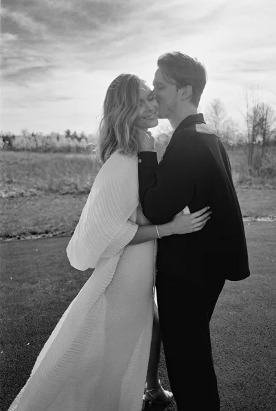 Wedding kiss portrait of bride and groom embrace, his forehead kiss as her pleated bridal cape drapes in a cloudy field with trees