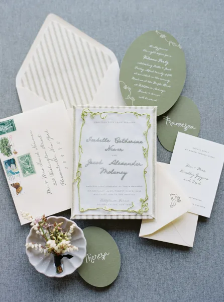 Invitation suite wedding invitation set with calligraphy, oval cards, ribbon trim, greenery border, and vintage stamps on gray fabric flat lay