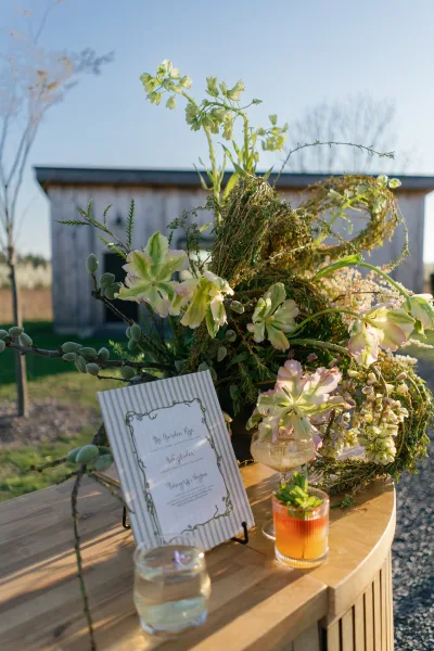 Wedding welcome table with wedding welcome table decor, floral arrangement and greenery wreath on wood table by a barn under blue sky