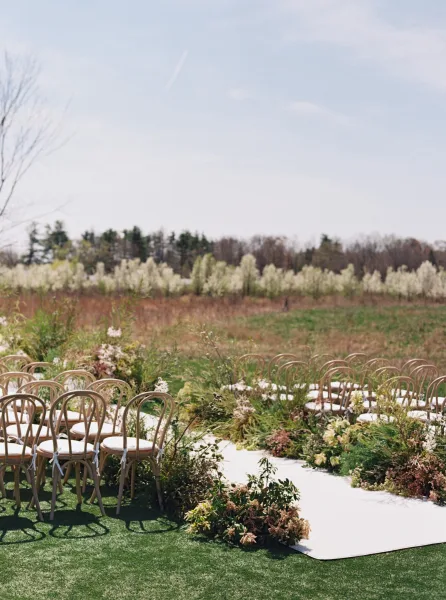 Ceremony aisle decor with a white aisle runner flanked by meadow florals and greenery, wood chairs with ribbons on a sunny lawn field