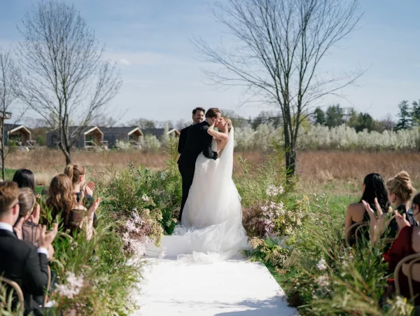Ceremony kiss as bride and groom kiss on a white aisle runner, her veil flowing between grounded florals in an outdoor field setting