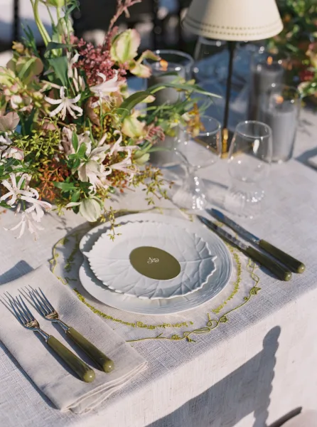 Reception tablescape with wedding place setting, scalloped plates, embroidered napkin, floral centerpiece, and taper candles in sunlit garden greenery