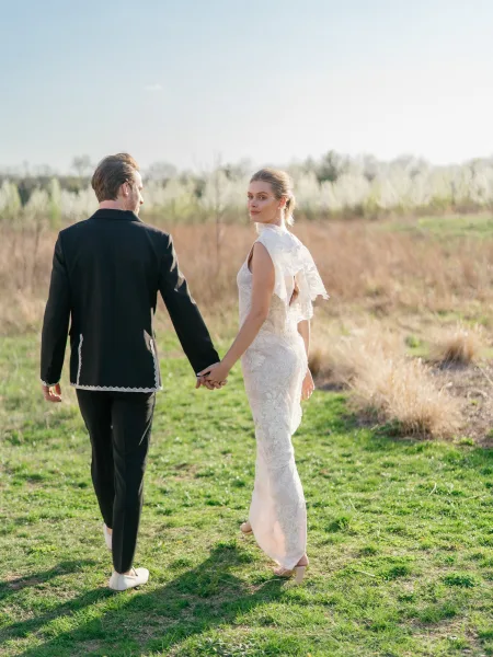 Couple portrait of bride and groom walking away holding hands in a sunny meadow, bride looking back in a lace wedding dress