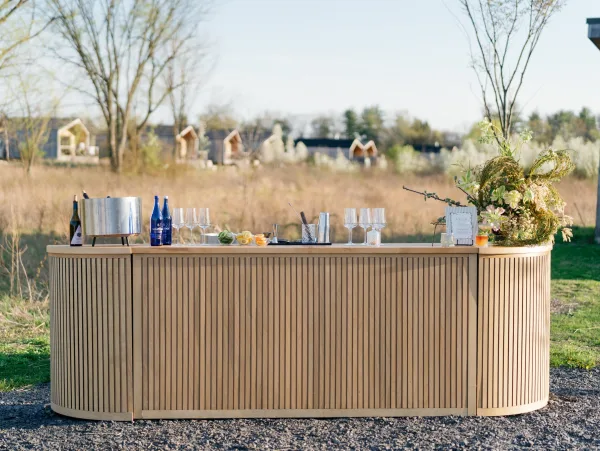 Wedding bar setup on a wood slat bar with bottles, glasses, citrus garnish, and candle accents in a meadow under blue sky