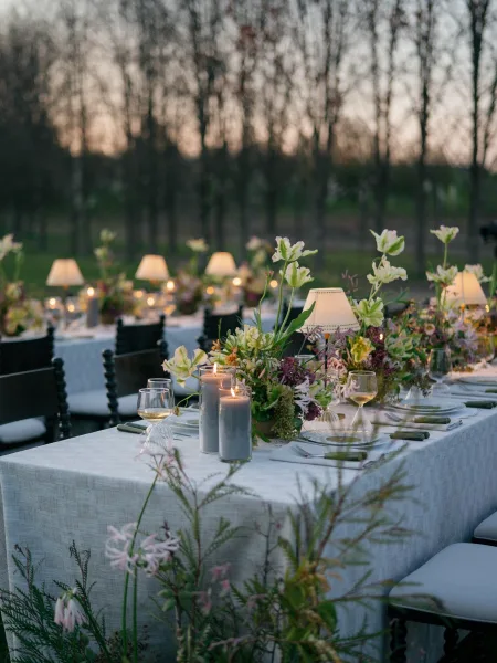 Reception tablescape with an outdoor wedding dinner table set in a garden at dusk, featuring floral centerpieces, taper candles, and table lamps