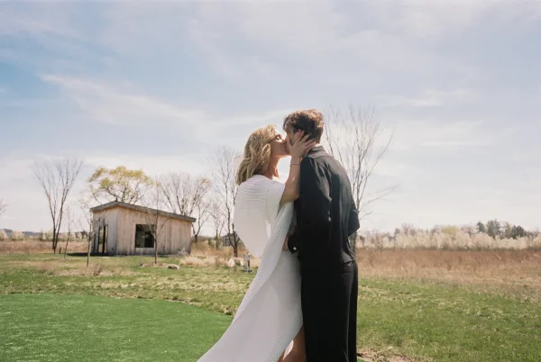 Wedding kiss as bride in long-sleeve gown and veil holds groom’s face, backlit in a grassy field with bare trees and rustic building
