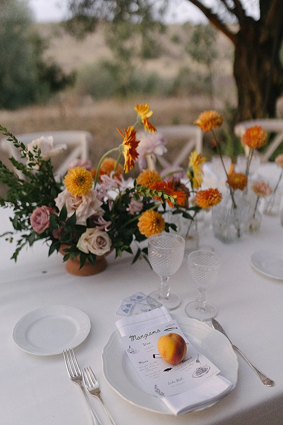 Reception tablescape with wildflower centerpiece, roses and orange blooms on a white tablecloth, patterned goblets, fruit place setting, countryside view