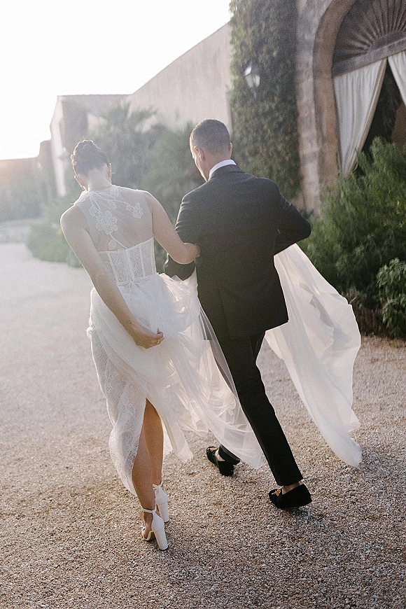 Newlywed exit as bride and groom walk away, bride holding her dress train on a sunlit gravel path by a stucco archway
