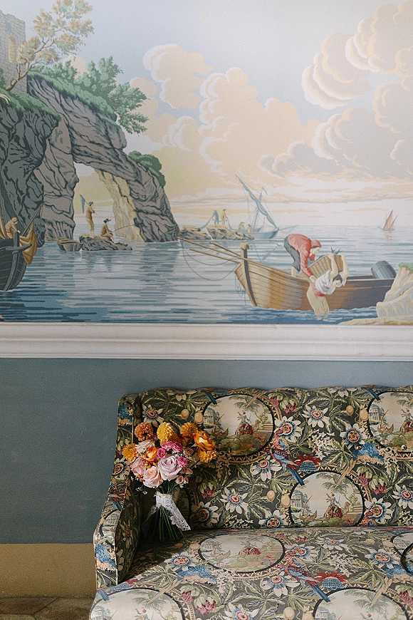 Bridal bouquet with garden rose bouquet in pink and yellow, wrapped in lace ribbon, resting on a patterned sofa before an ocean mural backdrop