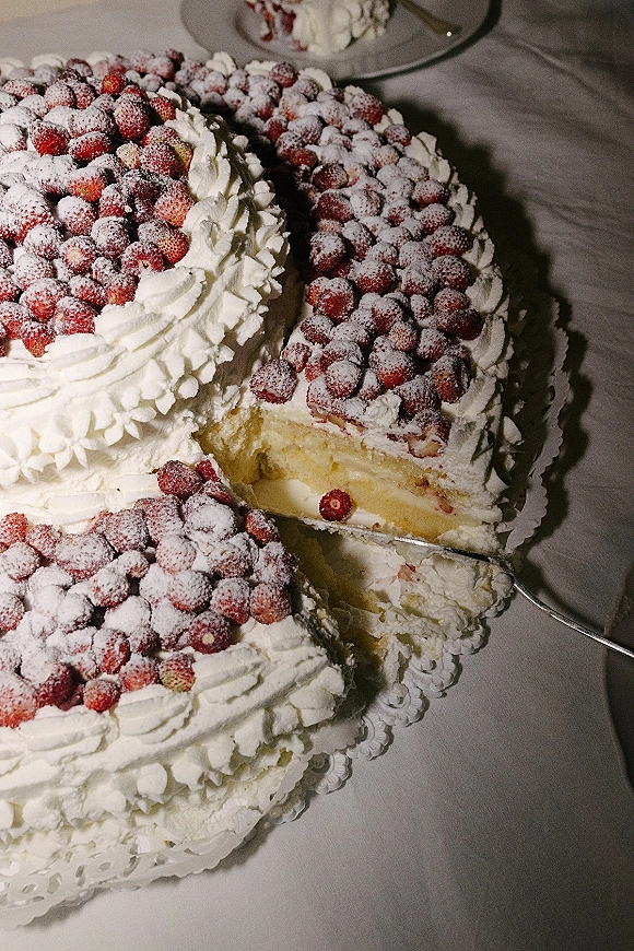 Wedding cake with whipped cream frosting and a strawberry wedding cake slice being served on dessert plates atop a tablecloth