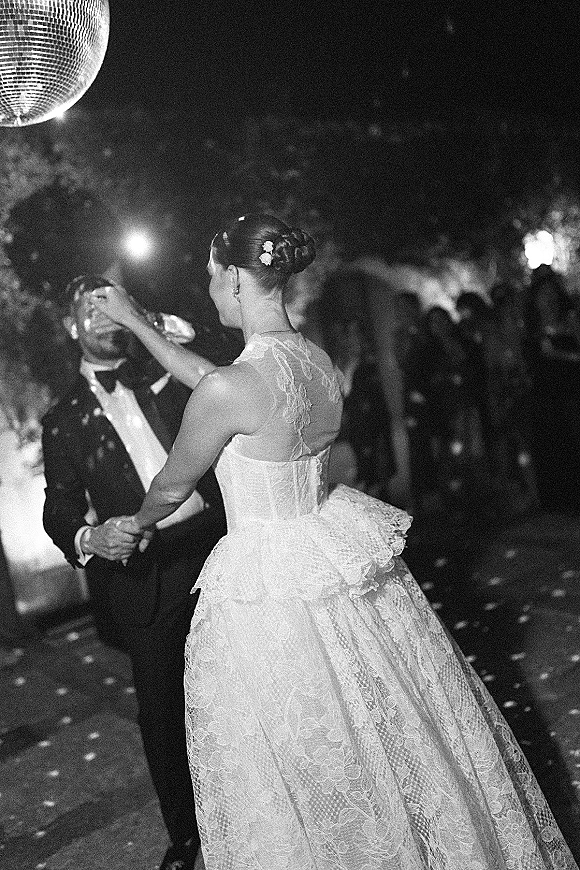 First dance with bride and groom dancing under a disco ball, her lace tulle gown and floral updo lit by spotlights outdoors at night