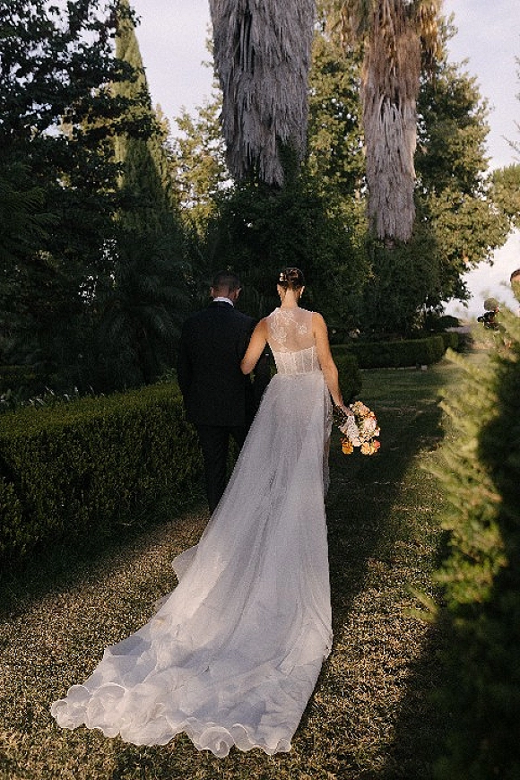 Couple portrait of bride and groom walking away, her lace-back gown with long train and bouquet along a tree-lined garden path