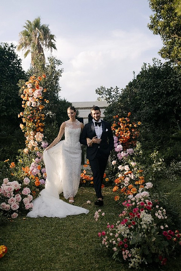 Newlywed portrait of bride and groom walking under a floral arch, her veil flowing as they cross a lush garden lawn with palm tree backdrop