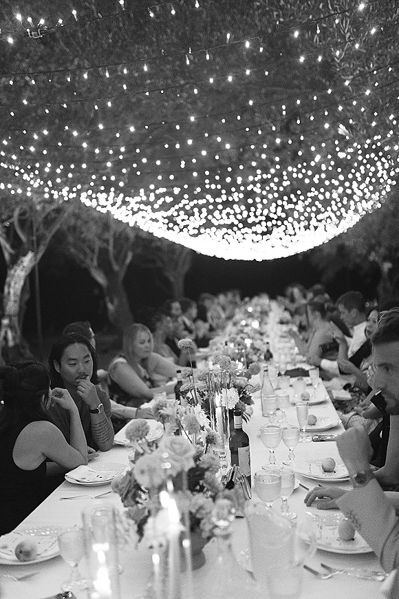 Reception dinner under a string light canopy, long banquet table set with floral centerpieces and candles, guests seated among trees at night