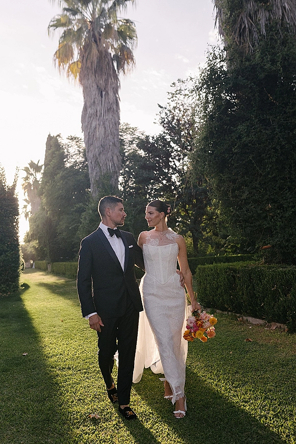 Couple portrait of bride and groom walking on a sunlit garden lawn with palm trees, bride holding a colorful bouquet and veil flowing