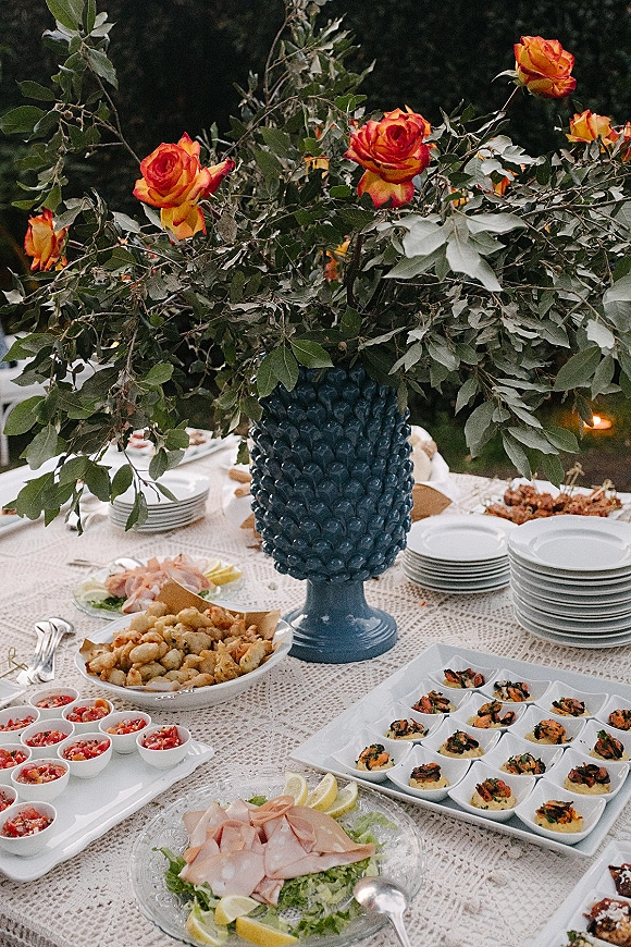 Wedding catering display with cocktail hour appetizers in cups, orange roses and greenery centerpiece on lace tablecloth in a garden setting