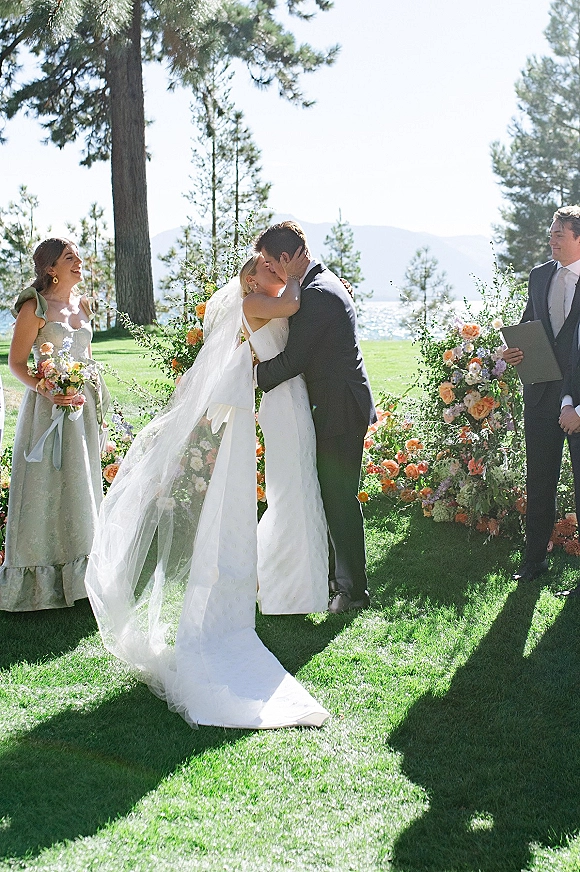 Wedding kiss as the bride and groom kiss under a floral arch, veil blowing, with lake and mountains behind at the ceremony