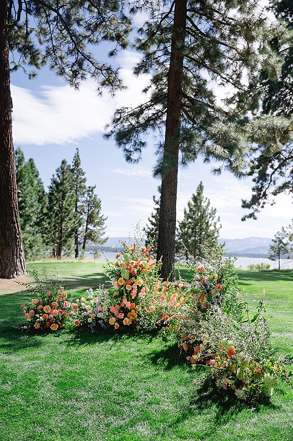 Ceremony aisle florals with grounded roses and meadow wildflowers lining a lawn aisle, set against pine trees, a lakeside mountain view