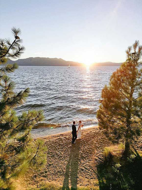 Couple portrait of bride and groom holding hands on a sandy beach at sunset, bride with bouquet and veil, mountains and sailboat behind