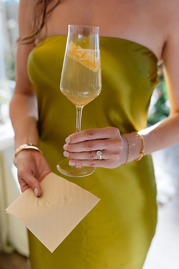 Bridal toast as bride holding champagne in a flute with citrus garnish, rings and gold bracelets visible in soft indoor light by a doorway