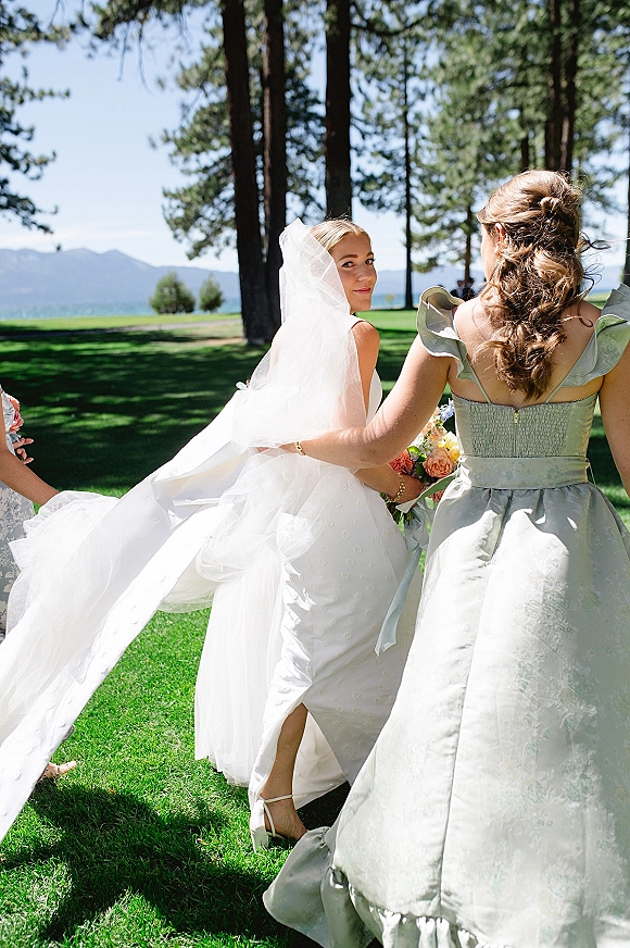 Bridal party moment as bride with bridesmaids walks with veil flowing, bride holding bouquet on lakeside lawn with pines and mountains