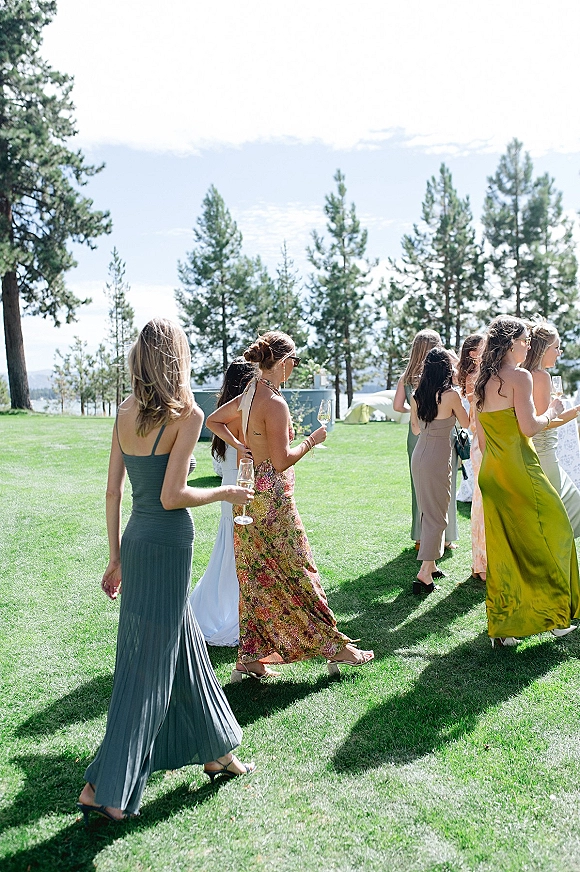 Wedding cocktail hour with wedding guest mingling as bridesmaids in formal gowns hold champagne flutes on a lakeside lawn by pines and mountains