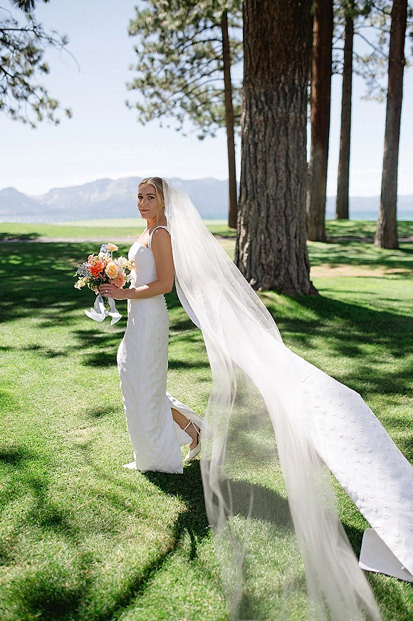 Bridal portrait of a bride holding bouquet with a long cathedral veil blowing in wind on a lawn by pine trees, lake and mountains