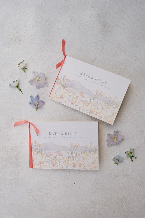 Wedding invitations with a watercolor wedding invitation design, tied with coral ribbon and pressed flowers on a light textured surface