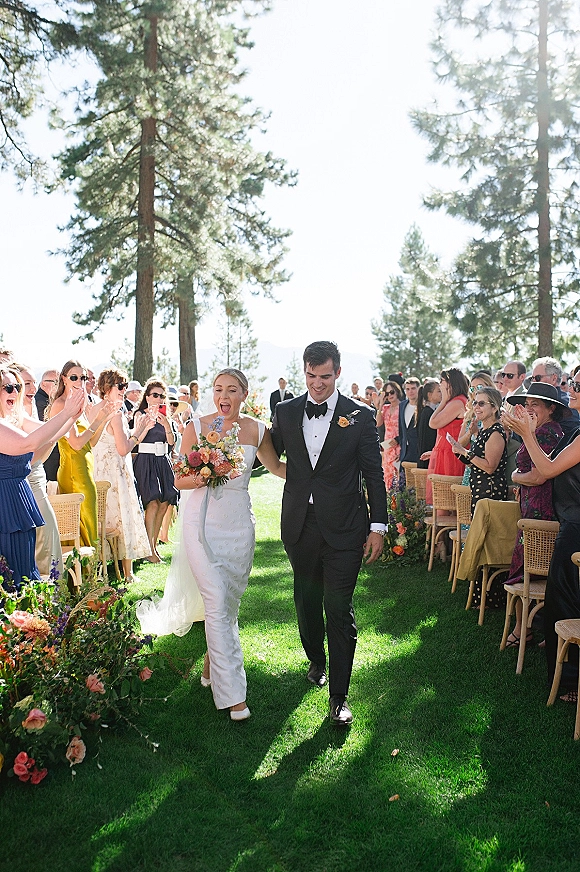 Wedding recessional as bride and groom walk the aisle, her bouquet raised, guests cheering on a sunny lawn beneath tall pine trees