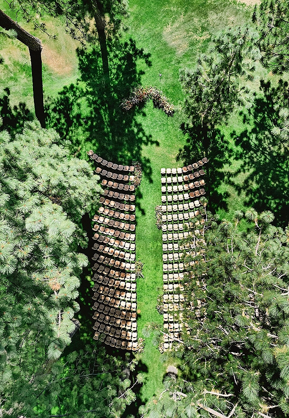 Outdoor ceremony setup with wood chairs in two blocks facing a floral arch and aisle floral clusters on a grass lawn under pine trees