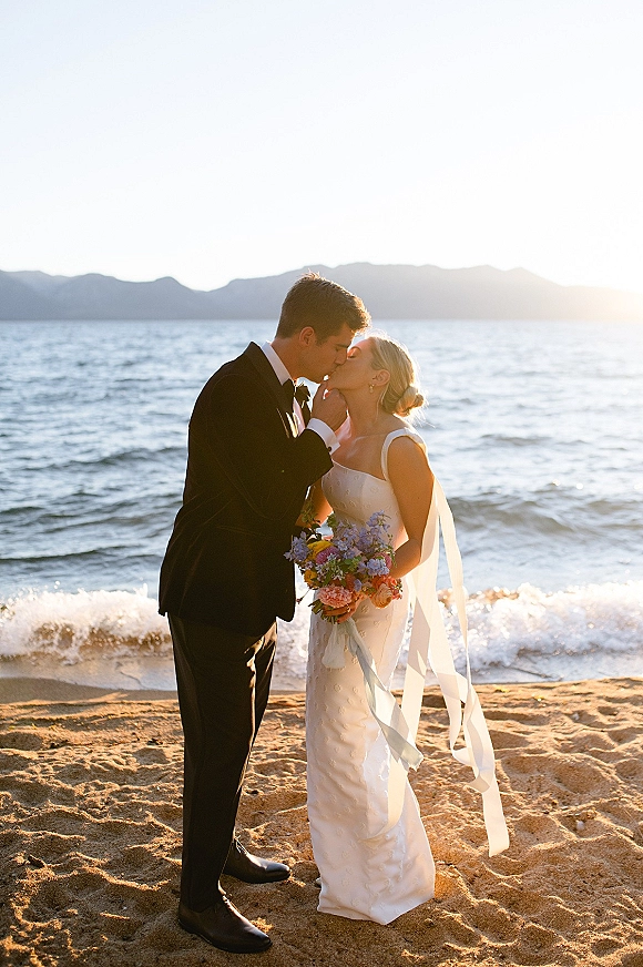 Wedding kiss portrait of bride and groom kissing at sunset on the beach, veil blowing in wind as she holds a colorful bouquet with ribbons