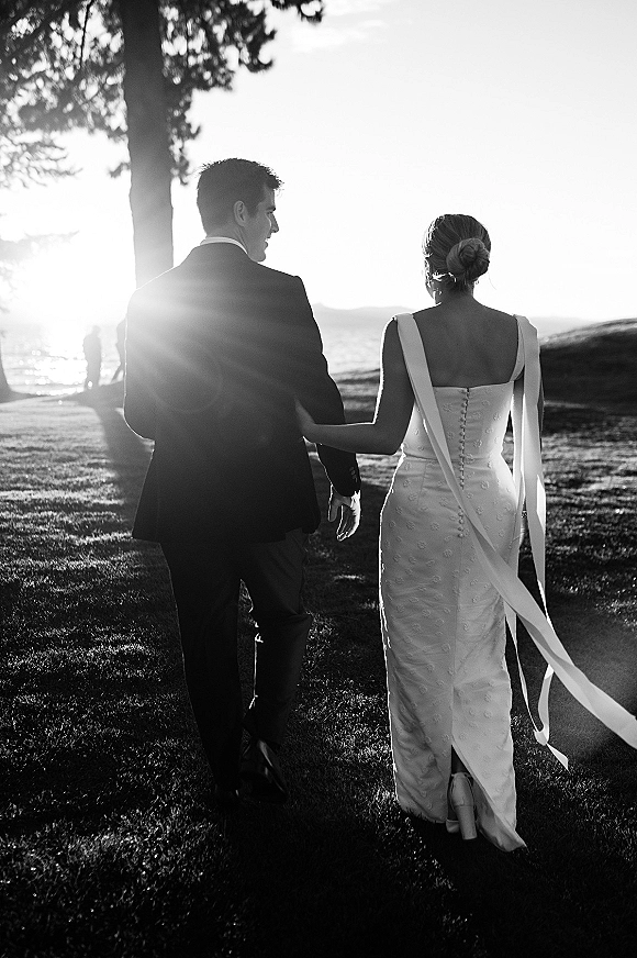 Couple portrait of bride and groom walking away holding hands, sunlit on a lawn by the ocean horizon, her button-back dress visible