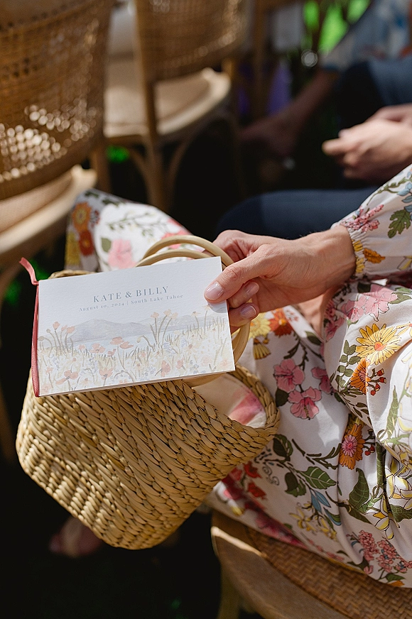 Wedding ceremony program with ceremony program design tucked in a woven straw basket with ribbon, near wicker chairs and seated guests