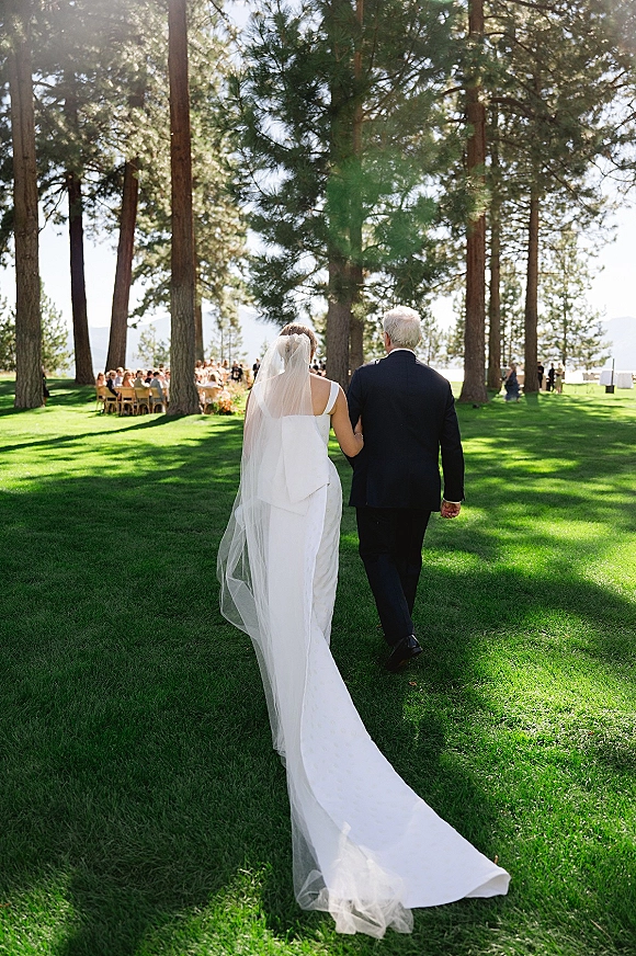 Processional moment as bride walking down aisle from behind, long veil flowing, with guests seated at sunny lakeside pine venue