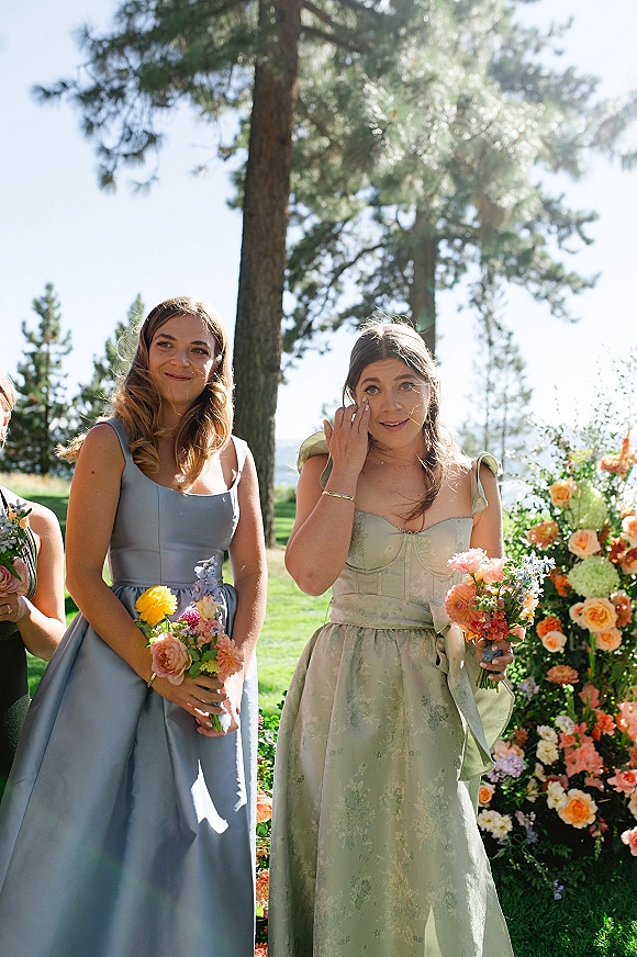 Bridesmaid portrait of bridesmaids holding bouquets in mismatched dresses, standing on a sunny lawn with pine trees and blue sky behind