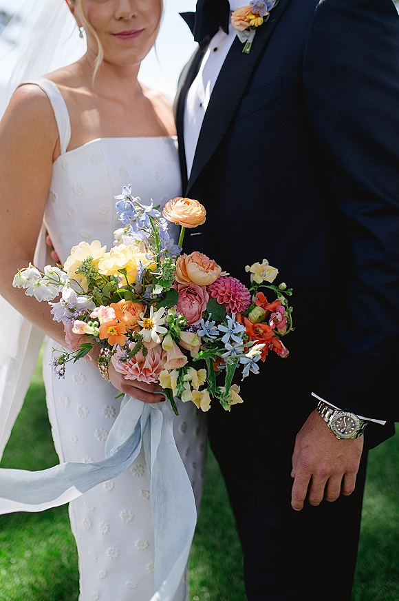 Couple portrait of bride and groom holding a colorful bouquet, her veil draped over a simple dress on a grassy lawn under sky
