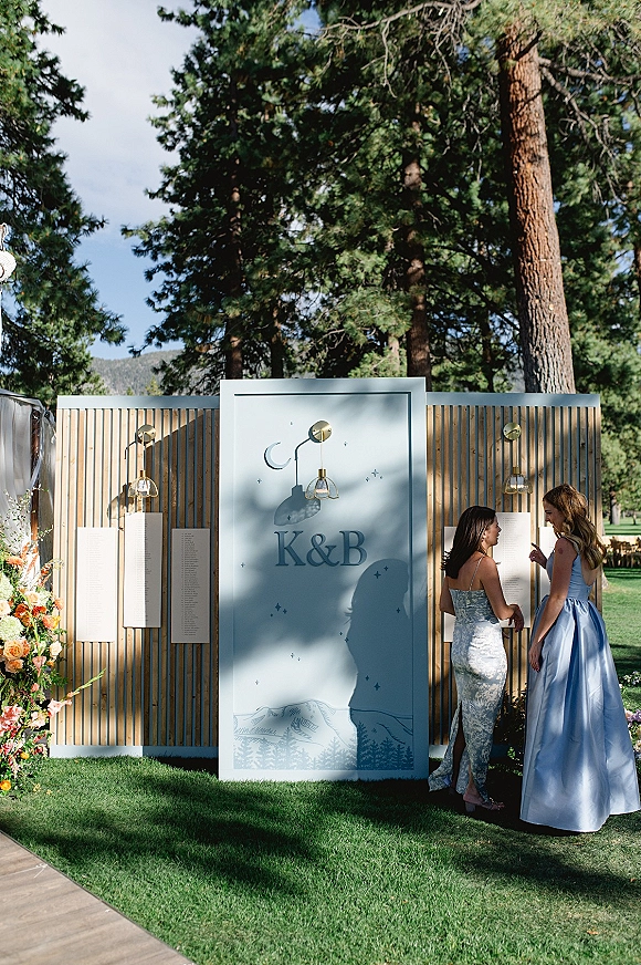 Wedding seating chart escort card wall with monogram panel, sconces, floral arrangement and draped fabric on a wood slat backdrop outdoors