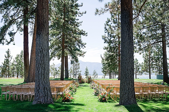Outdoor ceremony setup with wooden chairs and colorful floral aisle arrangements on grass, set by a pine forest lake with mountains
