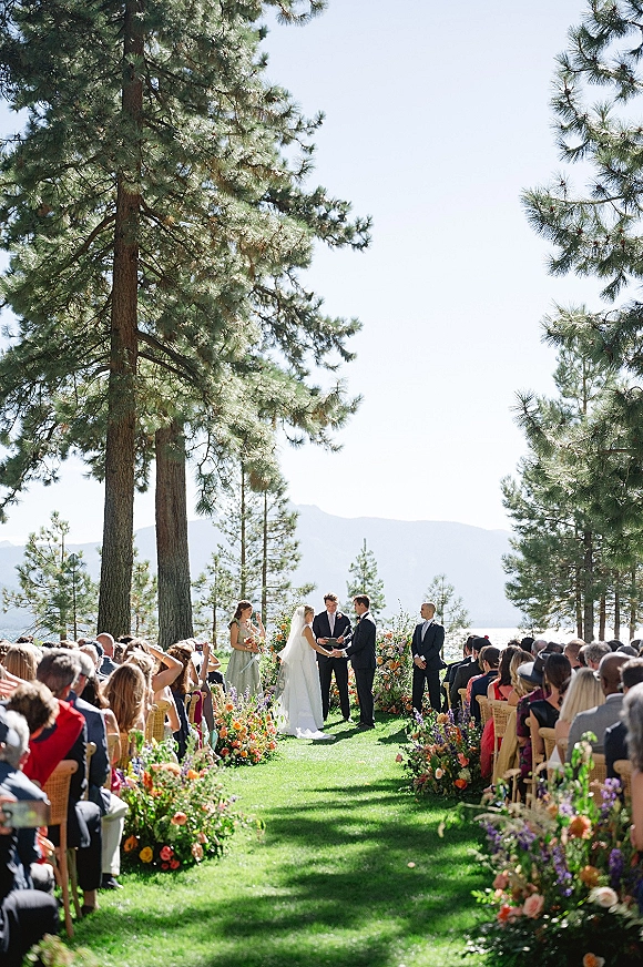 Wedding ceremony with bride and groom at altar, floral-lined aisle and arch, by a lakeside lawn with pine trees and mountains behind